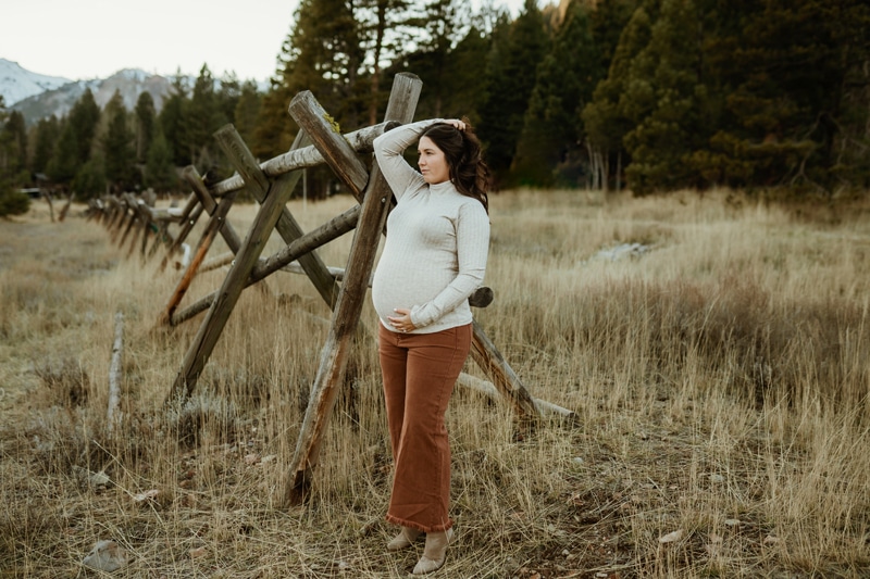 A mom to be in brown pants and a white sweater holds her bump and hair back while standing in a pasture against a fence