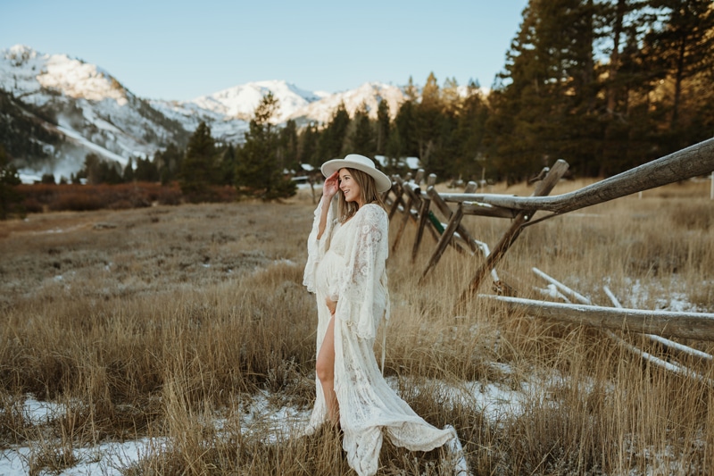 A mom to be in a large white hat and white lace maternity gown stands in a snowy field at sunset with a hand on her bump