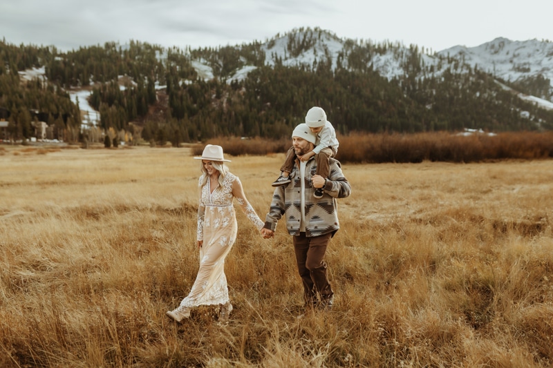 A mom and dad walk through a pasture holding hands while their toddler sits on dad's shoulders during christmas in lake tahoe