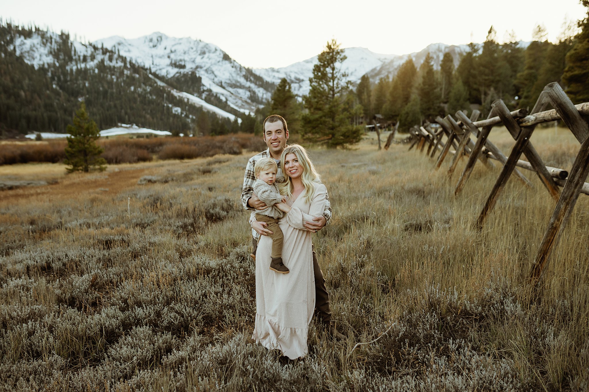 A mom and dad stand in a pasture by a wooden fence holding their toddler son at sunset during a stay at a family friendly hotels lake tahoe