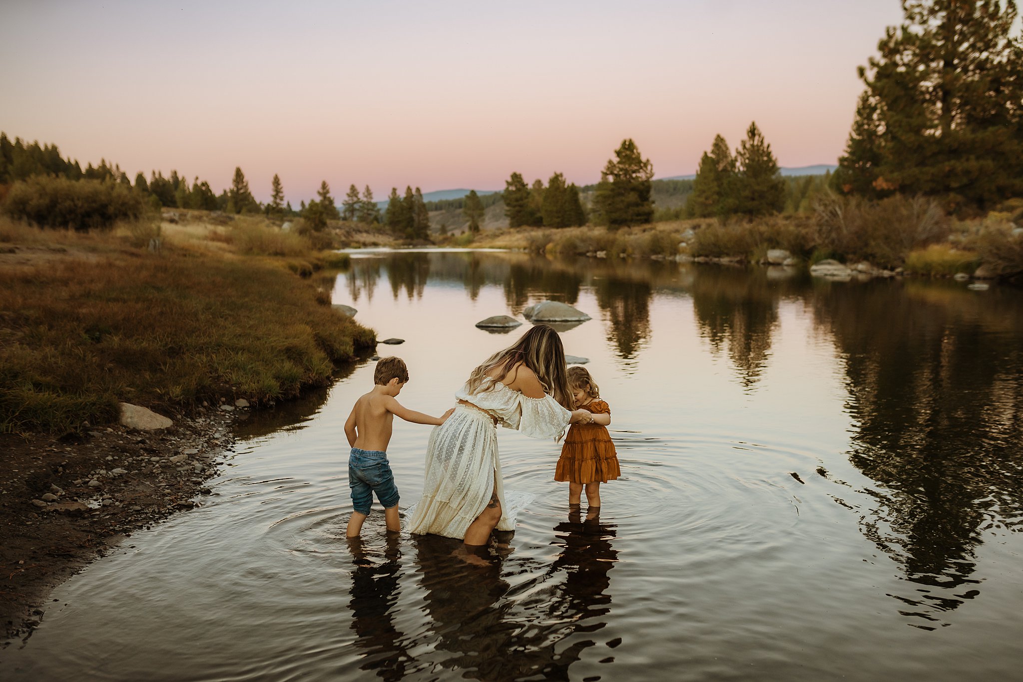 A mother in a white dress plays with her toddler daughter and son in a shallow river at sunset after visiting reno toy stores
