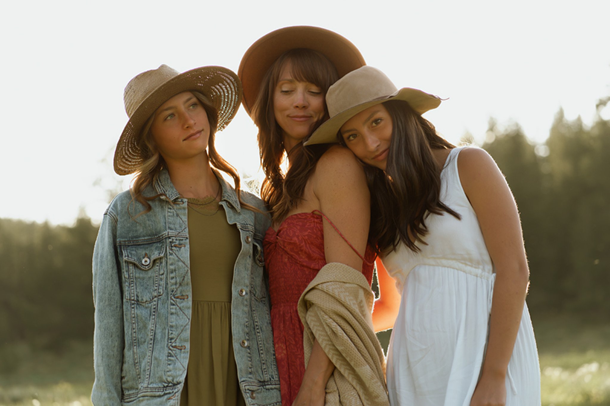 Two teenage daughter lean onto their mother while standing in a field at sunset wearing hats